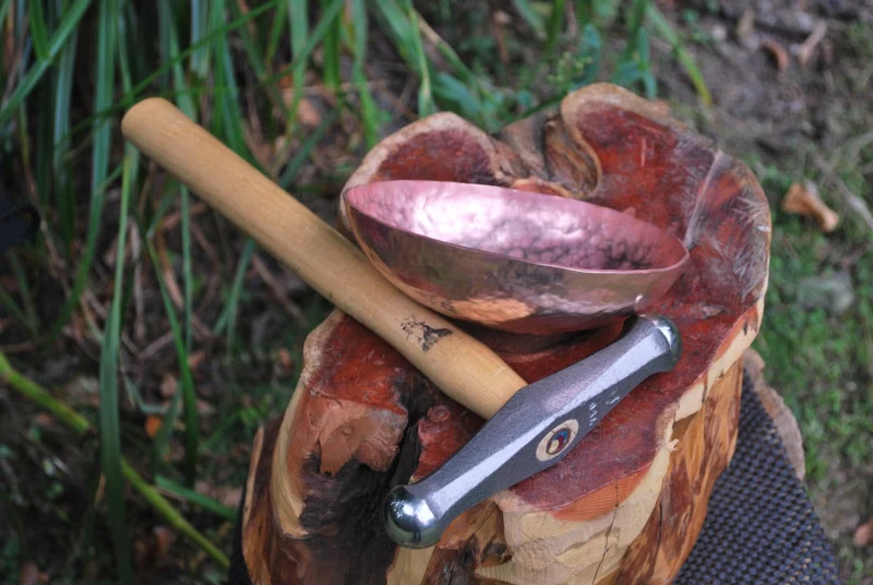 A hand-hammered copper bowl and a forging hammer resting on a tree stump