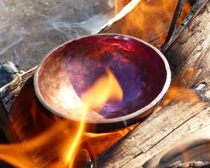 A hand-hammered metal sound bowl glowing copper and violet as it rests in a wood fire